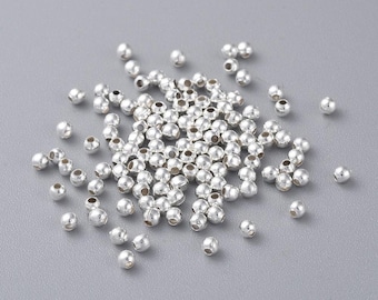 May include: A large pile of silver metal beads, scattered on a gray surface. The beads are round and smooth, and they are all the same size.