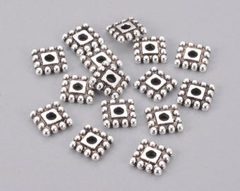 May include: Silver-colored square beads with a central hole and a beaded border. The beads are scattered on a gray surface. These decorative beads are suitable for jewelry making and craft projects.