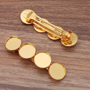 May include: Two gold-coloured barrettes with circular settings. One barrette has four settings, and the other has three. The barrettes are made of shiny metal and are designed to hold small items or embellishments. They are displayed on a wooden surface.