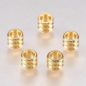 May include: Five gold-coloured, cylindrical beads with a ridged design. Each bead has a central hole and a polished, metallic finish. The beads are arranged on a light grey surface, showcasing their reflective quality.