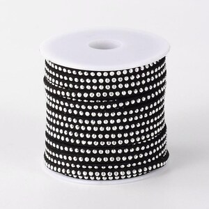 Puede incluir: Un carrete de cordón de ante negro con diamantes de imitación plateados. El cordón está enrollado alrededor de un carrete de plástico blanco.