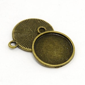 Op de afbeelding: Een paar ronde, bronzen metalen hanger-schalen. De schalen zijn ontworpen om een rond, plat stuk glas of ander materiaal te bevatten. De bovenste schaal heeft een decoratief, zonnestralenpatroon en een kleine lus om een ketting of koord aan te bevestigen.