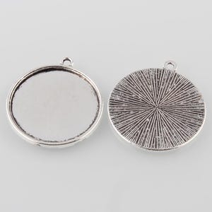 Op de afbeelding: Twee zilverkleurige ronde hangerblanks. Eén heeft een plat, verzonken oppervlak en de andere heeft een radiaal patroon op de achterkant. Elk heeft een kleine lus om aan een ketting of koord te bevestigen. Ideaal voor het maken van sieraden.