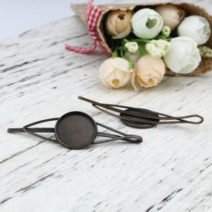 Op de afbeelding: Twee antieke goudkleurige haarclips met ronde, lege metalen insteekjes. De clips liggen op een wit houten oppervlak.
