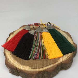 May include: Five colorful tassels with silver tops, displayed on a wooden slice. The tassels are red, black, rainbow, yellow, and green.