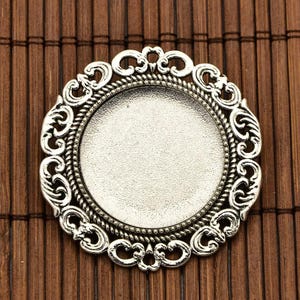 May include: Silver-toned metal pendant with a round, ornate frame and a blank circular space in the centre. The frame has a detailed, intricate design with swirls and curves.