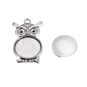 Op de afbeelding: Zilveren uilbedel met een ronde, blanco, glazen cabochon in het midden. De bedel is ontworpen om te gebruiken voor het maken van sieraden of knutselen.