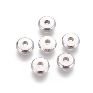Può includere: Un insieme di sei piccole perline metalliche circolari color argento. Ogni perlina presenta una superficie liscia e lucida con un bordo esterno rialzato e un foro centrale, progettata per essere infilata. Le perline sono disposte in gruppo.