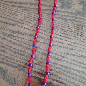 May include: Red cord necklace with blue beads and a wooden cross pendant. The necklace is knotted and features small blue beads spaced along the red cord. The cross is a light brown color and has a simple, classic design.