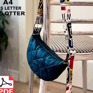 Op de afbeelding: Een tealblauwe, gewatteerde heuptas met een wit en rood gepatroneerde band. De band heeft tekst erop, waaronder "WILM" en "NO". De heuptas hangt aan een witte stoel met een gepatroneerde zitting.