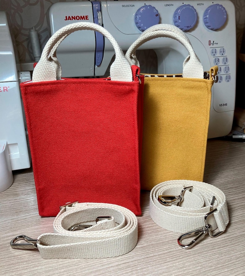 May include: Two small canvas tote bags with white straps and a detachable strap. The bag on the left is red and the bag on the right is yellow.