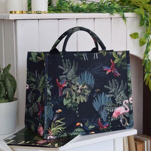Op de afbeelding: Een zwarte tote bag met een tropische print met daarop toekans, papegaaien en roze flamingo's op een donkergroene achtergrond.