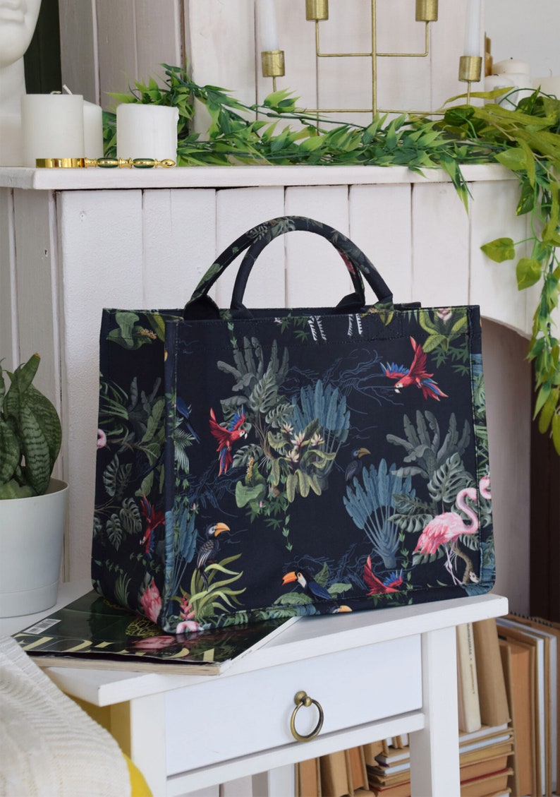 Op de afbeelding: Een zwarte tote bag met een tropische print met daarop toekans, papegaaien en roze flamingo's op een donkergroene achtergrond.