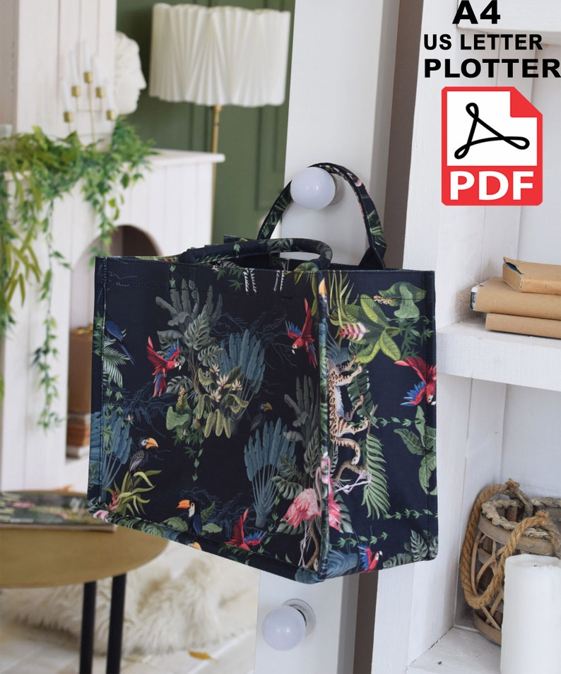 Op de afbeelding: Een zwarte tote bag met een tropische print met daarop toekans, papegaaien, flamingo's en junglebegroeiing. De tas hangt aan een wit rek. De tekst "A4 US LETTER PLOTTER PDF" is zichtbaar in de rechterbovenhoek van de afbeelding.