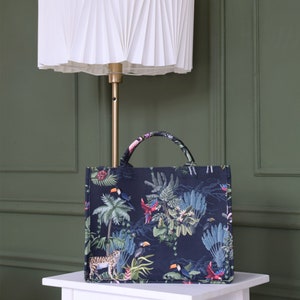 Op de afbeelding: Een zwarte tote bag met een tropische print met daarop toekans, papegaaien en een jaguar. De tas staat voor een witte geplooide lampenkap.