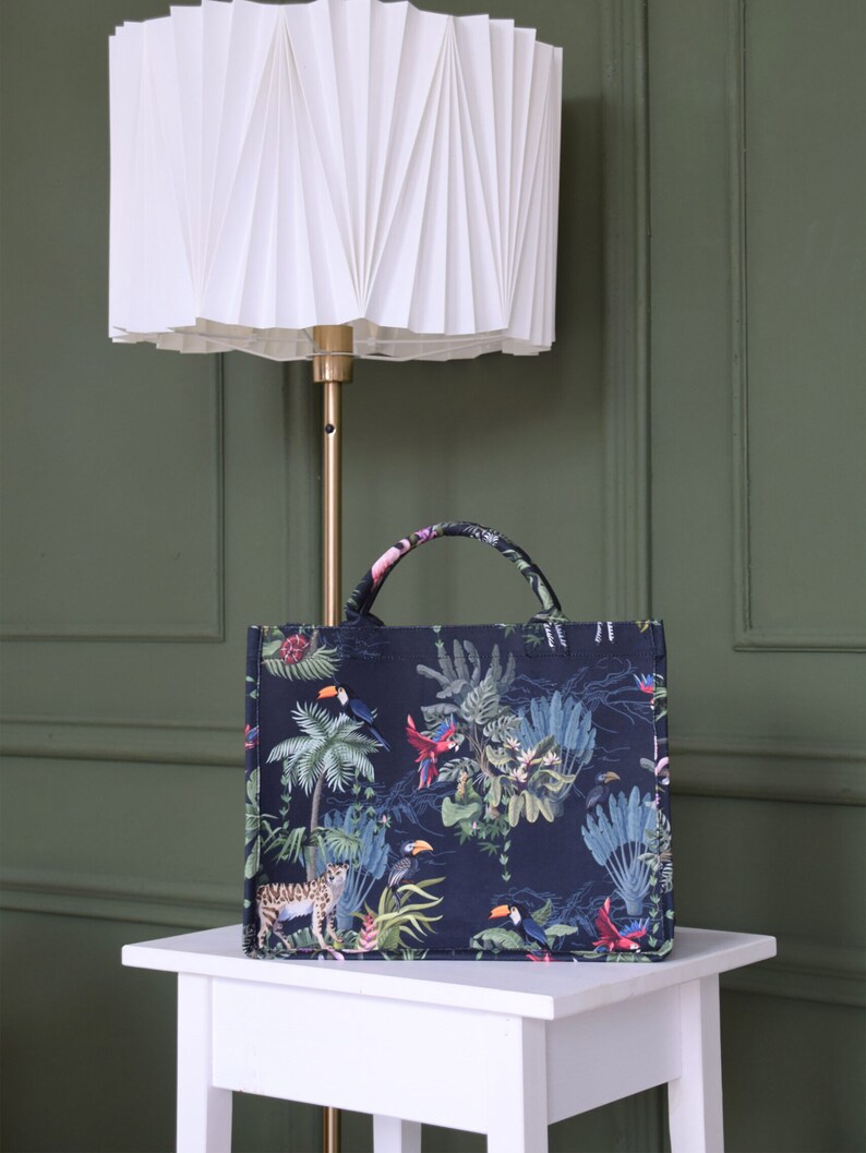 Op de afbeelding: Een zwarte tote bag met een tropische print met daarop toekans, papegaaien en een jaguar. De tas staat voor een witte geplooide lampenkap.