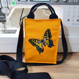 May include: A small yellow crossbody bag with a black strap and a butterfly embroidered on the front. The butterfly is yellow, black, blue, and red.