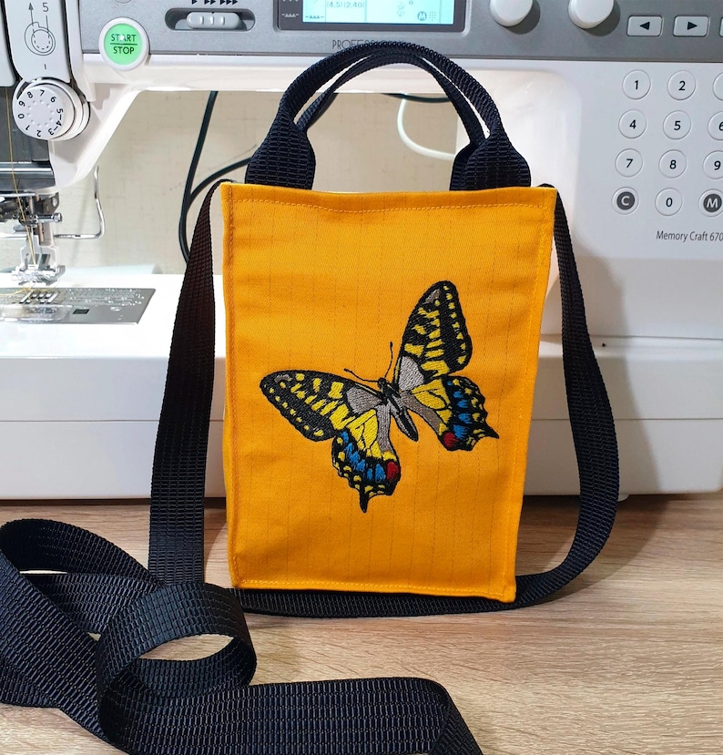 May include: A small yellow crossbody bag with a black strap and a butterfly embroidered on the front. The butterfly is yellow, black, blue, and red.