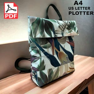 以下が含まれることがあります： 緑、茶色、青の色合いの熱帯の葉のプリントが施された白いキャンバスのバックパック。バックパックはフラップ留めと調節可能な黒いストラップが付いています。画像の上部に「A4 US LETTER PLOTTER」というテキストが黒で印刷されています。