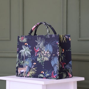 Op de afbeelding: Een zwarte tote bag met een tropische print met daarop toekans, papegaaien en palmbomen. De tas heeft twee handvatten en een ritssluiting.