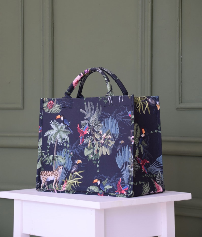 Op de afbeelding: Een zwarte tote bag met een tropische print met daarop toekans, papegaaien en palmbomen. De tas heeft twee handvatten en een ritssluiting.