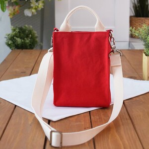 May include: A red canvas tote bag with a white shoulder strap and top handles. The bag has a zipper closure and a small pocket on the front.
