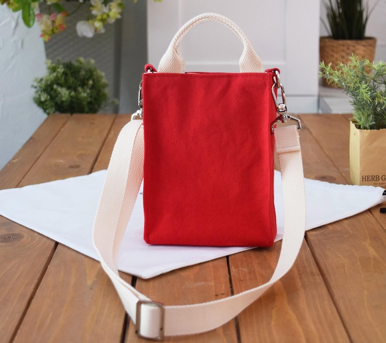 May include: A red canvas tote bag with a white shoulder strap and top handles. The bag has a zipper closure and a small pocket on the front.