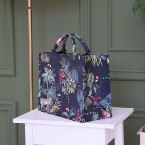 Op de afbeelding: Een zwarte tote bag met een tropische print met toekans, papegaaien en junglebegroeiing. De tas heeft &eacute;&eacute;n bovenste handvat.