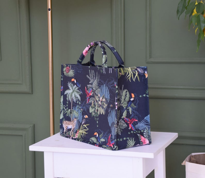 Op de afbeelding: Een zwarte tote bag met een tropische print met toekans, papegaaien en junglebegroeiing. De tas heeft &eacute;&eacute;n bovenste handvat.