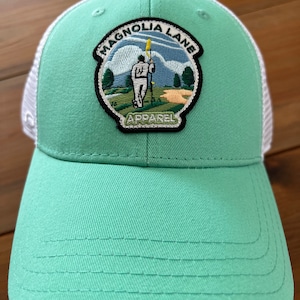 May include: A green and white trucker hat with a patch that says "Magnolia Lane Apparel" and features a golfer on a green.
