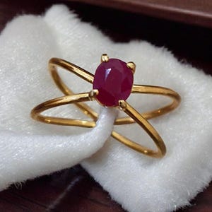 Op de afbeelding: Een gouden ring met een rode ovale edelsteen in het midden. De ring heeft een dubbele band met de banden die elkaar kruisen.