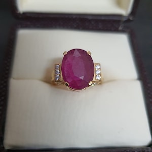 Puede incluir: Un anillo de oro con una gran piedra de rubí ovalada rodeada de diamantes más pequeños. El anillo está en una caja de anillo de terciopelo blanco.