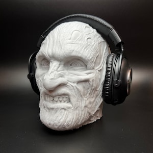 May include: A white 3D printed sculpture of a face wearing black headphones. The face has a detailed, textured surface and a menacing expression.