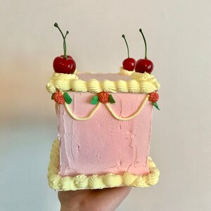 May include: A square pink cake with yellow frosting and red cherries on top. The cake is decorated with green leaves and white frosting.