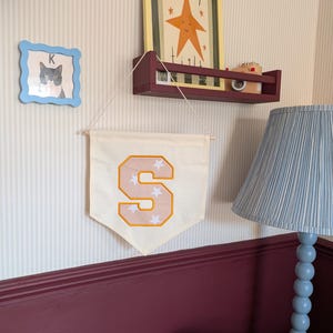 May include: A cream-coloured fabric wall banner featuring a large, centred letter S in a light peach hue with a yellow border. The letter is decorated with small white star patterns. The banner hangs from a thin string attached to a wooden dowel.