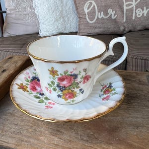 May include: A white teacup and saucer with a floral pattern in shades of pink, yellow, blue, and green. The cup and saucer have a gold trim.