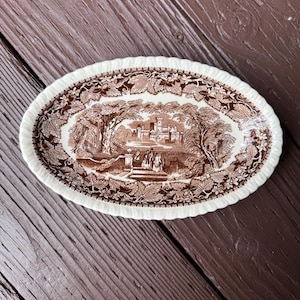 May include: An oval ceramic dish with a cream-colored base and a brown transferware pattern. The design features a central landscape with a castle, trees, and figures on a terrace, surrounded by a border of detailed grape leaves and vines.