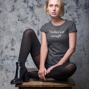 May include: A charcoal gray t-shirt with the text "Vanilla's not enough!" in white. The person is wearing black tights and black boots. The background is a mottled gray and white.