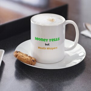 May include: A white ceramic mug with the text "Money yells but Wealth whispers" in green and orange. The mug sits on a white saucer, next to a chocolate chip cookie. The mug is filled with a foamy beverage.
