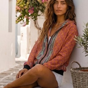 Puede incluir: Una chaqueta con cremallera y capucha que presenta un patrón repetitivo en forma de abanico en tonos naranja, rojo y verde azulado, con puños y dobladillo de canalé negro. El texto "कला KALA SARI" aparece impreso en negro en la esquina superior derecha.