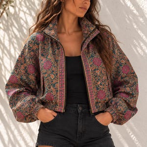 Puede incluir: Una chaqueta corta con cremallera sobre una base de color verde azulado oscuro con un vibrante estampado floral y de cachemira en tonos magenta, naranja y crema. La chaqueta tiene cuello alto, mangas largas con puños elásticos y dobladillo.
