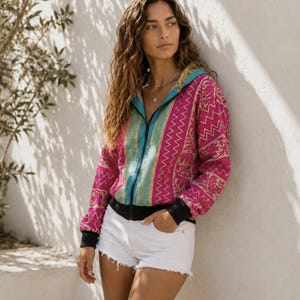 Puede incluir: Una chaqueta de manga larga con cremallera, confeccionada en una tela estampada de color magenta y dorado con motivos de zigzag y flores. La prenda incluye un forro interior turquesa, cremallera frontal negra y puños y dobladillo acanalados negros.