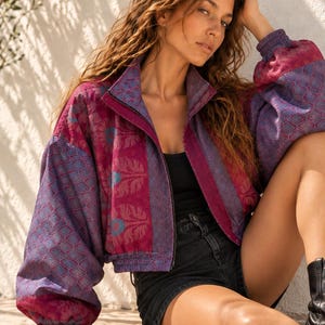 Pode incluir: Um casaco curto com fecho de correr apresentando um padrão floral vibrante em roxo e magenta, com gola alta e mangas compridas. O casaco é usado sobre uma camisola preta sem mangas e calções pretos, exibindo um tecido texturizado com punhos elásticos.