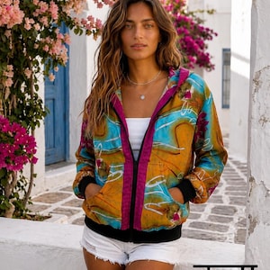 Può includere: Una giacca con cappuccio e cerniera caratterizzata da un motivo astratto nei toni del giallo senape, turchese e magenta. Il capo presenta polsini e fascia in vita neri, una chiusura lampo color magenta e due tasche frontali. Il testo Kala Sari è visibile.