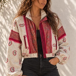 Pode incluir: Um casaco com fecho de correr em tom creme com um padrão paisley em vermelho e rosa escuro. O casaco tem gola alta, mangas compridas com punhos elásticos e painéis verticais vermelhos ao longo do fecho frontal. Usado sobre um top preto.