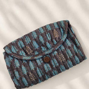 Puede incluir: Un estuche acolchado rectangular de tela gris carbón oscuro con un patrón repetitivo de motivos en forma de hoja en azul claro y dorado apagado. El estuche tiene una solapa redondeada asegurada por un botón de madera marrón en el centro.