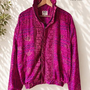 Op de afbeelding: Een levendig magenta jack met ritssluiting, een hoge kraag, lange mouwen met elastische boorden en een herhalend patroon van donkerblauwe en gouden paisley-motieven. Het binnenlabel bevat de tekst "कला" en "KALA SARI" in zwarte inkt.