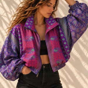 Puede incluir: Una chaqueta con cremallera que presenta un vibrante estampado de cachemira y flores en tonos violeta, rosa y azul cerceta. La chaqueta tiene cuello alto, mangas largas con puños elásticos y dobladillo elástico. Se lleva sobre un top corto negro.