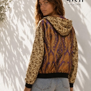 May include: A hooded jacket featuring a purple and orange paisley pattern on the back and a beige floral print on the sleeves and hood, with black ribbed cuffs and hem, paired with light blue denim shorts. The text KALA SARI is visible.