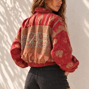 Op de afbeelding: Een rood jack met lange mouwen en een veelkleurig paisley- en geometrisch patroon, voorzien van een kraag en een elastische zoom, gecombineerd met een zwarte spijkerbroek. Het jack heeft een losse pasvorm en wordt over de zwarte short gedragen.